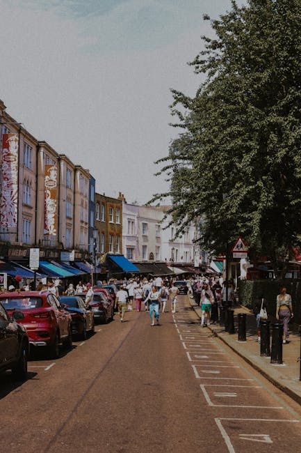 A narrow residential street in Notting Hill with a wet asphalt road marked by double yellow lines on each side. On the left, there is a weathered brick wall with an adjacent alleyway, some greenery ov