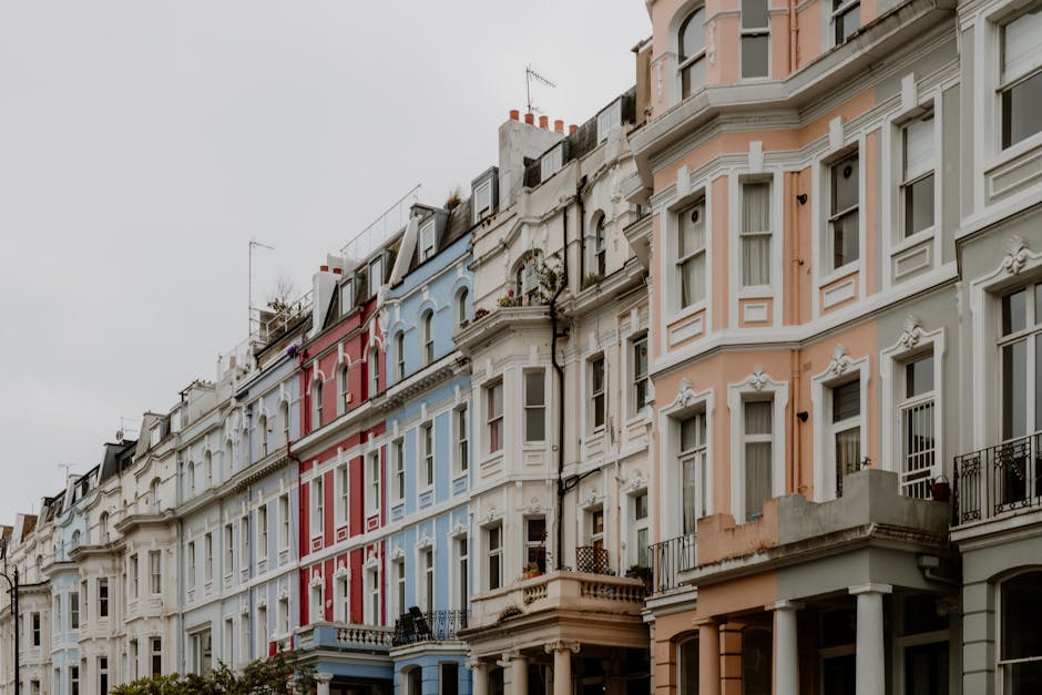 A street scene in Notting Hill showing a row of colourful terraced buildings with pastel facades, some with decorative balconies and large windows, under a partly cloudy sky. In the foreground, at the