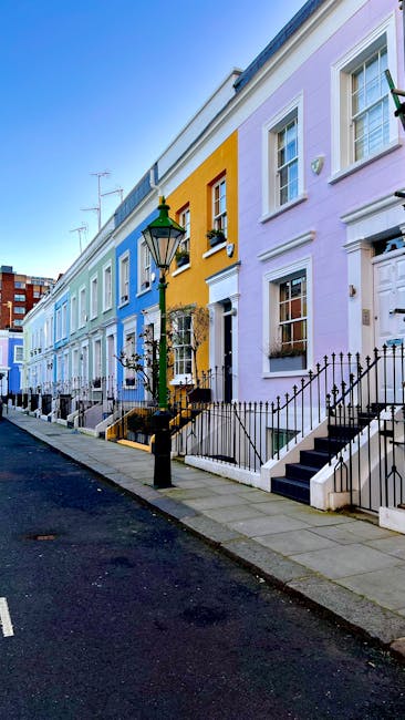 A row of brightly coloured terraced houses along a pavement in Notting Hill, with façades painted in pastel shades of blue, yellow, pink, and lavender. Each property has a small staircase leading up to a front door, some with black wrought iron railings and steps. A traditional street lamp stands near the pavement, with a green lantern-style top. The sky is clear and blue, indicating a sunny day. In the foreground, the black asphalt road is visible, with a white dashed line marking the parking area. The scene reflects a typical residential street in Portobello Road, with the visually appealing, vibrant architecture characteristic of the area. This picturesque setting supports visual context for house removals and moving services provided by Removal Company Notting Hill, highlighting the urban environment where professional furniture transport and home relocation activities take place.
