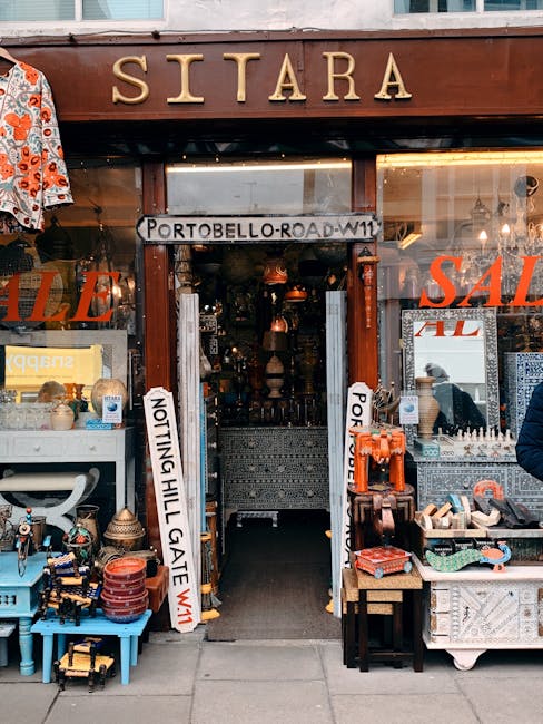 The storefront of a shop named 'Sitara' on Portobello Road W11 in Notting Hill features a wooden sign with the street name above the entrance. The exterior display includes various household items and decorative objects arranged on tables and shelves, with some items, including ceramics and textiles, partially visible through the glass windows. Two vertical signs are positioned on either side of the doorway, indicating the shop is not part of a cafe. Inside, the shop appears to be filled with furniture, lamps, and ornamental pieces, with warm lighting creating an inviting atmosphere. The surrounding pavement suggests the shop is situated on a busy street with pedestrians passing nearby. The shop front is part of a scene that can be associated with house removals or moving logistics, where careful packing and transport of various household goods are involved. This visual information from Removal Company Notting Hill supports understanding of home relocation and furniture transport processes.